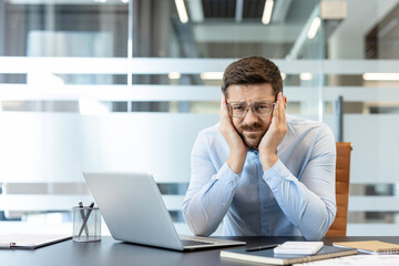 Stressed businessman experiencing an intense headache or migraine while sitting at a modern office...