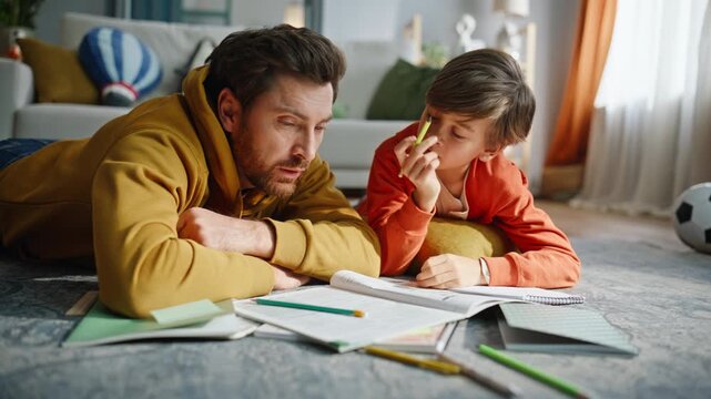 Father son solving school assignment looking textbook lying floor closeup. 