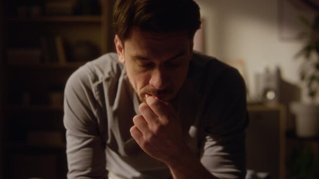 Tilt up shot of anxious adult man reading phone screen hesitating on texting back or doomscrolling news feed while sitting on bed in dimly lit bedroom
