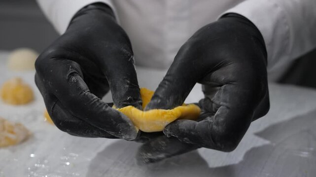 Baker hands cover cream filling with yellow rice dough to form cake in confectionery closeup. Confectioner prepares traditional Japanese cake with mango at kitchen countertop. Sweets eating