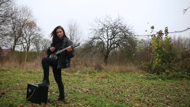 young female rock musician in a leather jacket plays an electric guitar outdoors