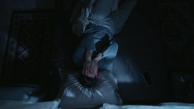 Top view of adult man lying in bed checking time on phone then rolling up in blanket trying to get to sleep during late night in bedroom