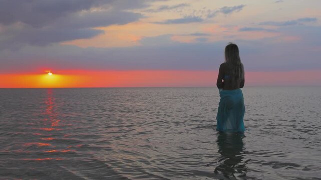 A girl in a blue swimsuit and a bright pareo posing against the background of a sunset in estuary with transparent water