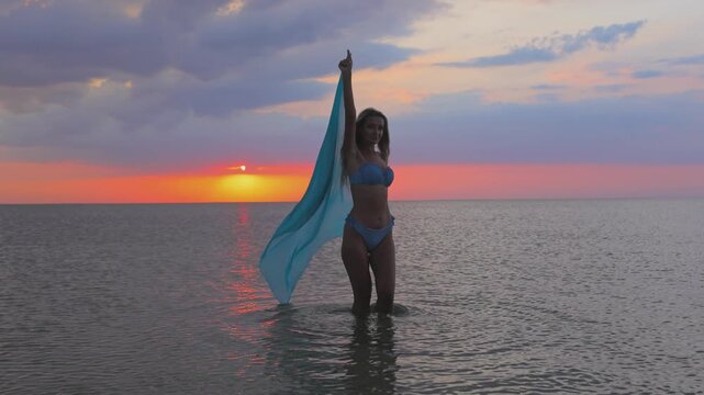 A girl in a blue swimsuit and a bright pareo posing against the background of a sunset in estuary with transparent water