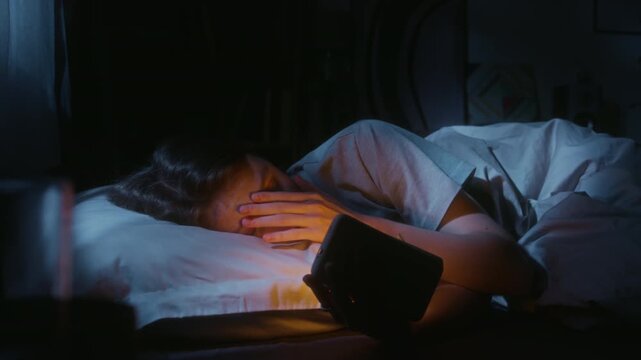 Young adult woman lying in bed scrolling through news on phone before bedtime reading stressful content late at night in dark bedroom