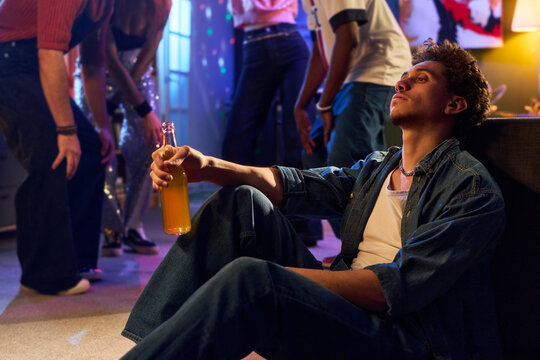 Young Black man sitting on floor holding bottle, appearing thoughtful while partygoers dancing in background, casual indoor party scene with diverse group of teenagers and young people