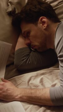 Vertical top view shot of adult man resting on bed covered with paperwork holding smartphone in hand while delaying work responsibilities in dim bedroom