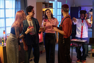 Fototapeta premium Group of multiethnic young people socializing and holding drinks at party while Black man DJing in background, everyone smiling and engaging in lively conversation indoors