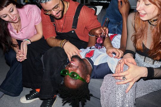 Group of young people including Black man wearing green sunglasses lying on couch laughing while multiethnic friends sitting around him smiling and holding drinks
