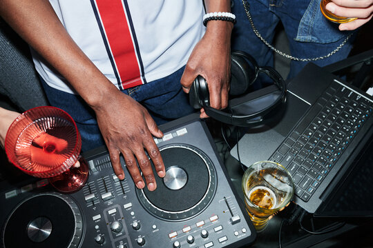 Black young man operating DJ controller with one hand while holding headphones, surrounded by drinks and laptop, other young people standing nearby holding beverages