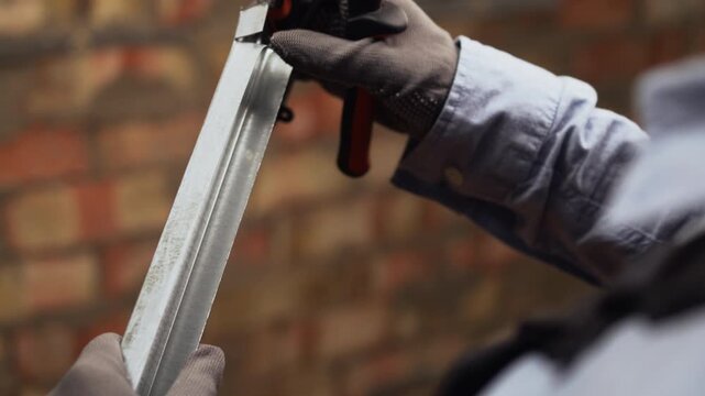 Close-up of female construction worker in overalls and gloves cuts iron profile for plasterboard with scissors. Builder cutting profiled metal with nippers.