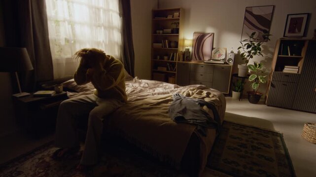 Wide angle shot of sunlit bedroom with young adult woman holding head coping with anxious thoughts then falling onto bed feeling fatigued