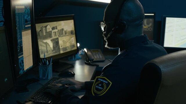 Cinematic side view shot of Black male dispatcher at police command center speaking to headset while watching computer monitors with data, backlit by monitor light during night shift, copy space