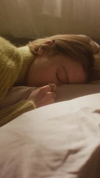 Vertical tilt up shot of drained young adult woman resting on pillow with eyes shut coping with overwhelming emotions in warm sunlit bedroom