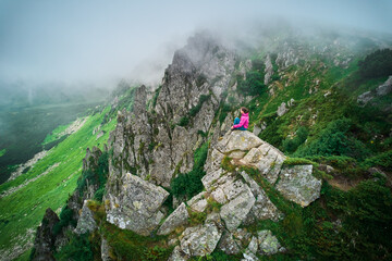 Hiker sits peacefully on large rock, high above lush, green valley, surrounded by jagged,...