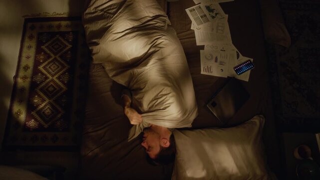 Top view of adult man lying in bed curled up under blanket suffering from depression and work procrastination while ignoring paperwork and incoming calls in low key lit bedroom