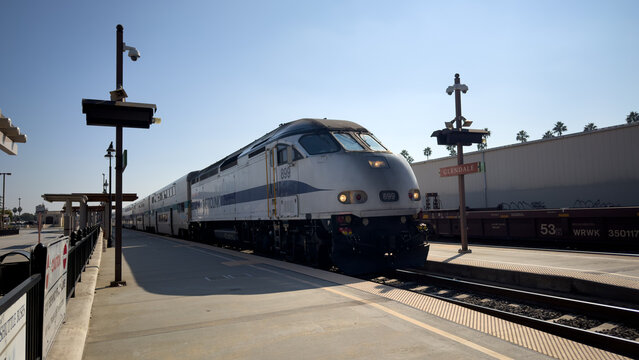 LOS ANGELES, CA, NOV 6, 2025: Metrolink locomotive arrives at Glendale station under clear blue skies, with a freight train visible on the adjacent track