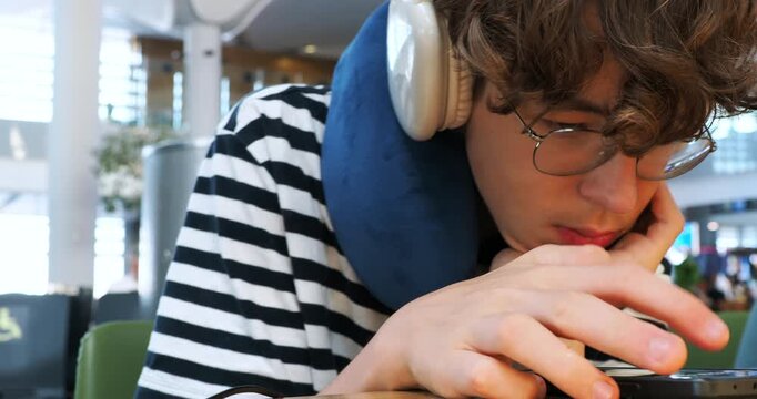 A tired young man with a neck pillow and headphones uses his smartphone while waiting at an airport. He looks sleepy and bored as he scrolls, killing time before his flight. Close-up shot