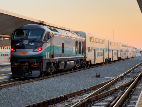 LOS ANGELES, CA, NOV 29, 2025: Metrolink locomotive train leads bi-level railway coaches at sunset on open tracks, warm golden sky, viewed from a station platform