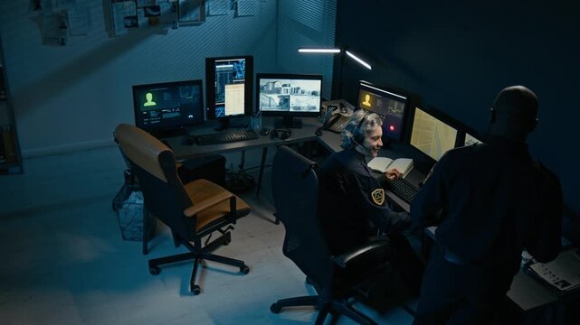 High angle view of dispatchers desk at police station with two people working and reviewing CCTV footage on multiple monitors during night shift, copy space
