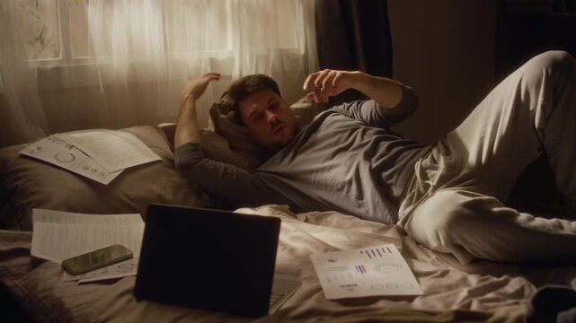 Middle aged man wearing pajamas lying on bed surrounded by documents struggling with procrastination problem while scrolling phone instead of working online on laptop