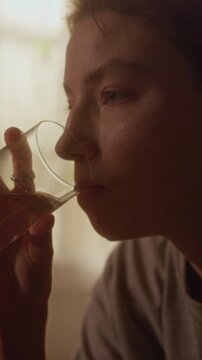 Vertical tilt up shot of young adult woman drinking from glass with quivering hands trying to soothe anxiety attack while sitting on bed in dimly lit bedroom
