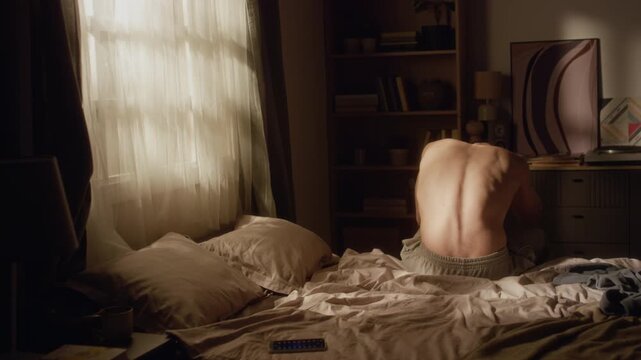 Rear view of shirtless adult man sitting on bed in morning throwing pillows with anger while feeling tense from stressful week