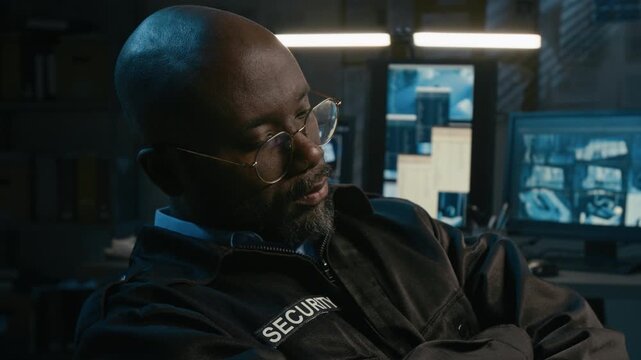 Tilt up shot of Black man working as security officer and falling asleep in control room during night shift with multiple surveillance computer monitors glowing in background