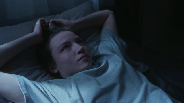 Medium shot of young adult woman lying in bed at night covering face with hands then sighing with exhaustion struggling to fall asleep