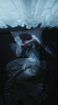 Vertical shot of young adult woman nervously rolling over in bed trying to get comfortable while struggling to fall asleep during night in bedroom