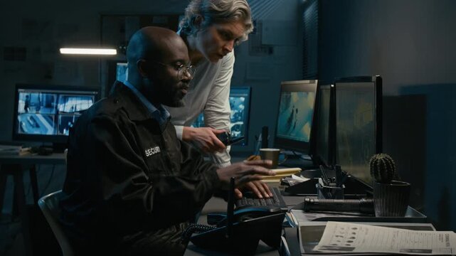 Side view shot of man and woman working together in security control room while watching surveillance footage and pointing at computer monitors during night shift, copy space