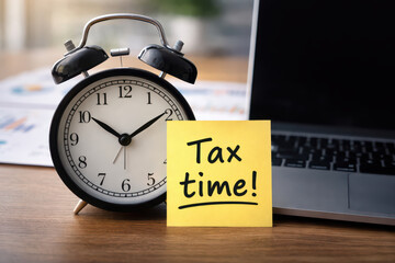 Tax time reminder note with classic alarm clock and laptop on wooden desk, symbolizing deadline and financial planning urgency