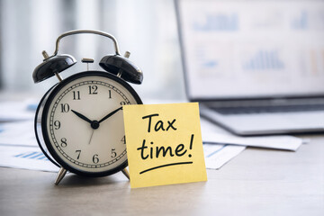 Tax time reminder note with classic alarm clock on office desk near laptop showing financial charts and documents, symbolizing deadline and urgency