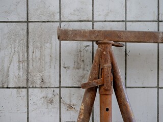 Close-up of a heavy-duty rusty steel construction tripod stand against a dirty white tiled wall,...