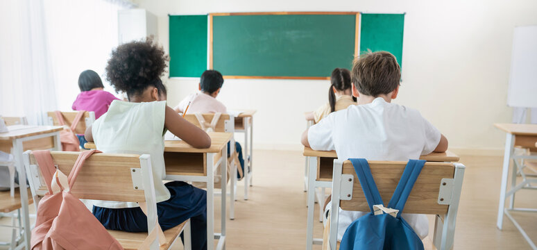 Portrait of little pupil writing at desk in classroom  elementary school. Student boy doing test from behide in primary school. Children writing in classroom. Education knowledge banner