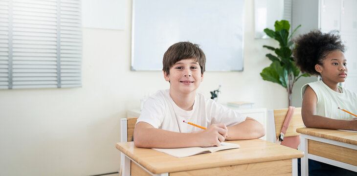 Portrait of little black caucasian pupil writing at desk in classroom at elementary school. Student boy study test in primary school. Children writing notes in classroom. Education knowledge banner