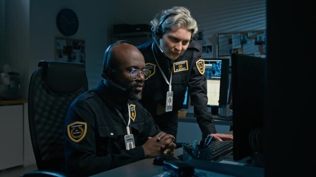 Medium shot of two police officers working together at station during night shift and using computer while pointing at monitor and coordinating operations, copy space