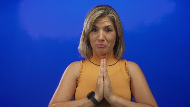 Blonde woman makes prayer gesture with clasped hands and looking upward against vivid blue studio wall; serenity.