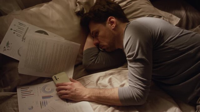 Top view of overwhelmed middle aged man lying on bed cluttered with documents holding phone in hand while struggling with procrastination in low key lit bedroom