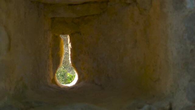 Keyhole shaped arrowslit in a medieval castle wall. Sunlight illuminating the interior of an ancient stone castle through a keyhole-shaped arrowslit, revealing a vibrant green landscape outside