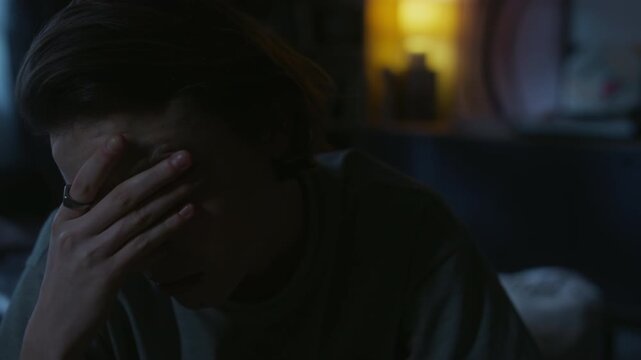 Medium shot of young adult woman pressing fingers to fatigue eyes remaining sleepless during night while sitting on bed in dark room