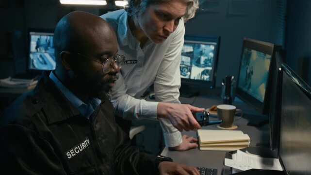 Tilt up shot of mature woman talking to African American man while coordinating security operation in control room and pointing at computer monitor showing CCTV feed working together at night