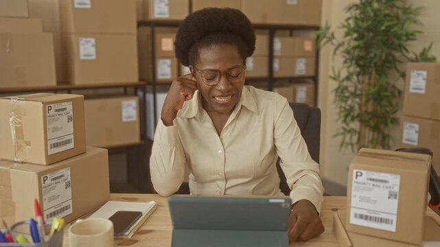 Woman business owner taps tablet and smiles while surrounded by stacked shipping boxes and a notebook on a desk in a building; entrepreneurial joy.