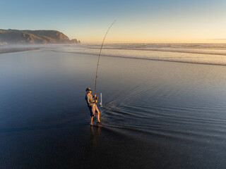A lone fisherman wades into the ocean at sunset, casting his line in search of a catch at the beach in Piha, Auckland, New Zealand. The sun reflects off the water.