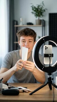 Teen male vlogger with acne recording skincare routine on smartphone with ring light. Influencer recommending blank tube cream for problem skin and pointing to face for UGC social media ad