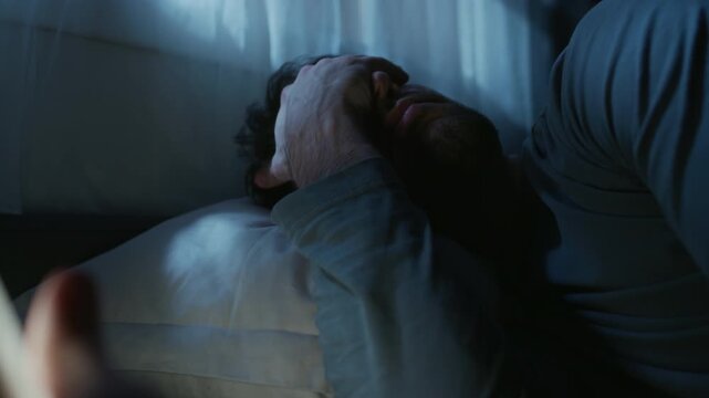 Close up on tired adult man browsing news feed on smartphone before sleep while reading social media at late night in dim bedroom