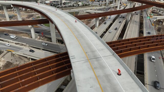 Elevated concrete highway ramp curve under construction with steel girders and orange safety cones spanning over a bustling interstate interchange 4k aerial drone video