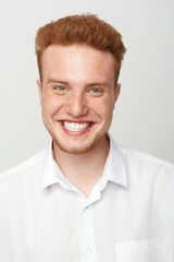 Obraz premium Smiling young man with red hair and freckles wears white shirt, looking at camera with bright happy expression. Studio portrait on light background, concept of joy and positive emotions.