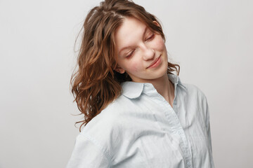 Obraz premium Young woman with wavy hair wearing white shirt looks down with gentle smile on light background. Concept of natural beauty, calmness, and soft emotions in portrait photography.