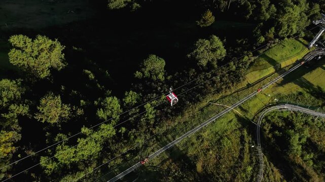 Aerial overhead view of cable car gondola and chair lift with people riding over pine forest with roller coaster tracks at Capivari amusement park in Campos do Jordao, Brazil. Static camera shot